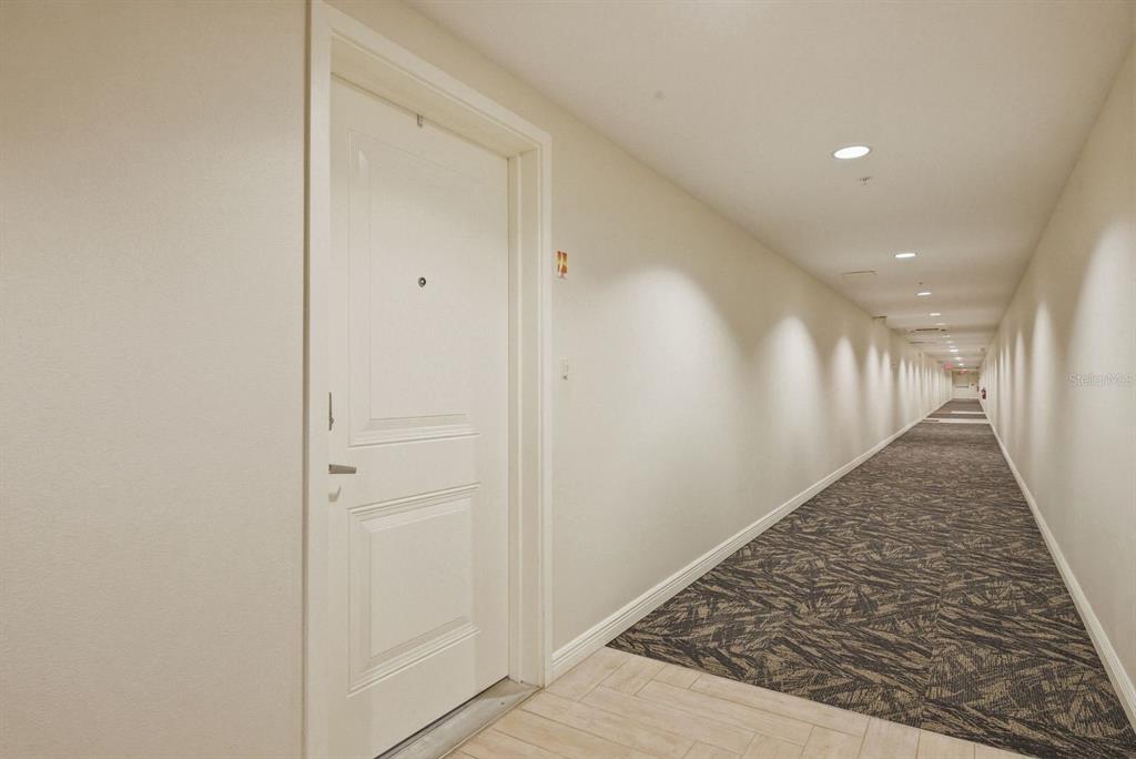 A long, narrow apartment hallway features a white door, beige walls, and patterned carpet flooring — 223 ISLAND WAY UNIT 2D, CLEARWATER BEACH, FL