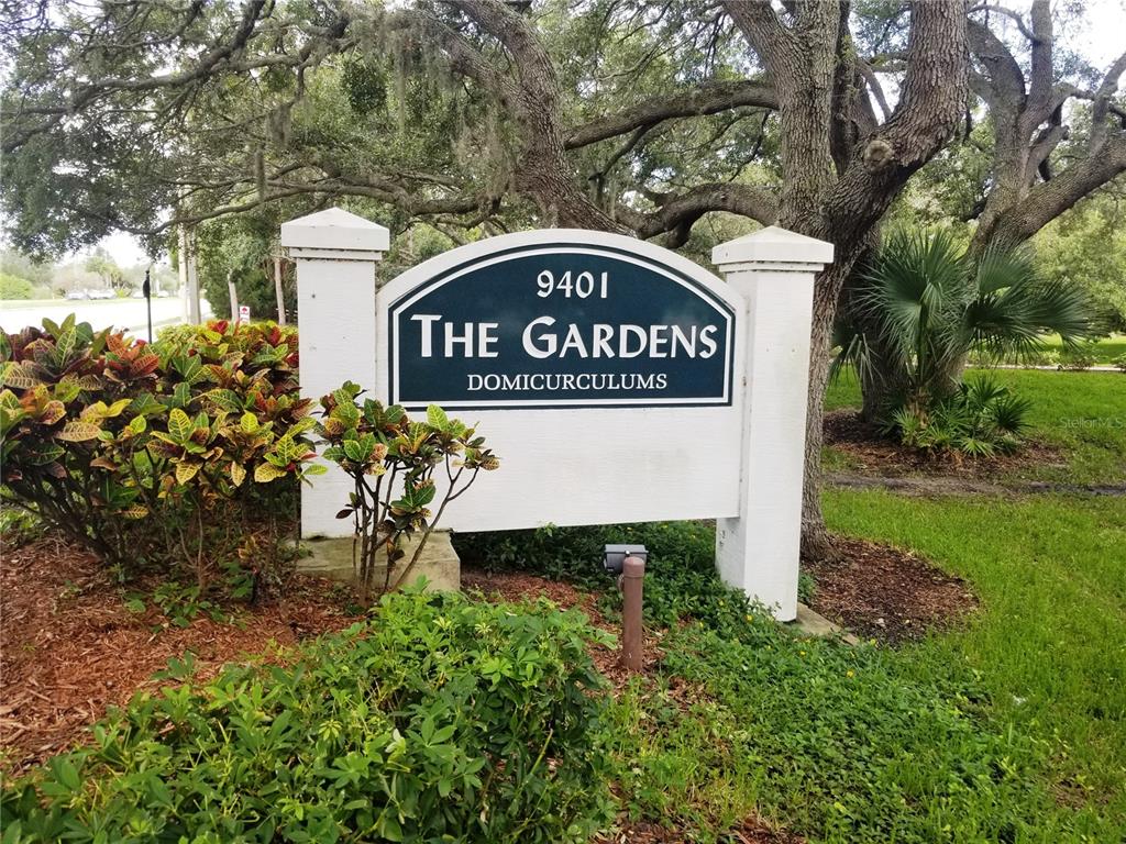 A sign for The Gardens stands in a landscaped area with trees and shrubs — 121 ASPEN CIRCLE UNIT 121, SEMINOLE, FL