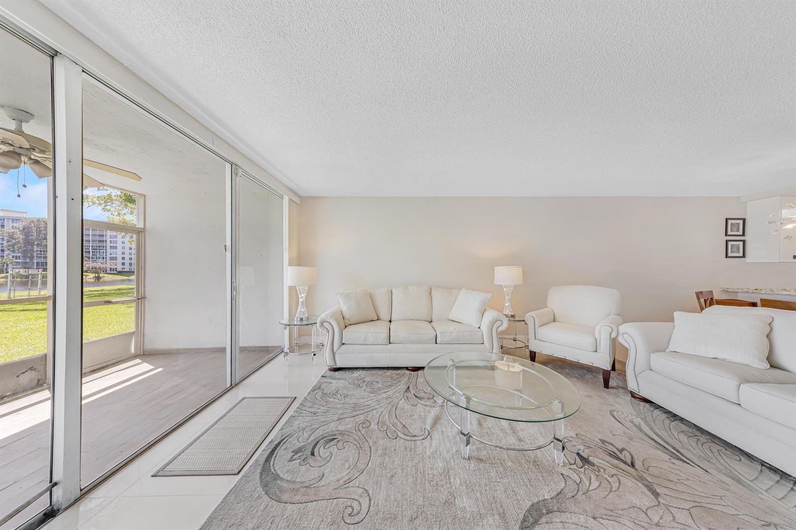 A living room features white furniture, a patterned area rug, and sliding glass doors to a patio — 3091 N Course Drive # 102, Pompano Beach FL 33069, POMPANO BEACH, FL
