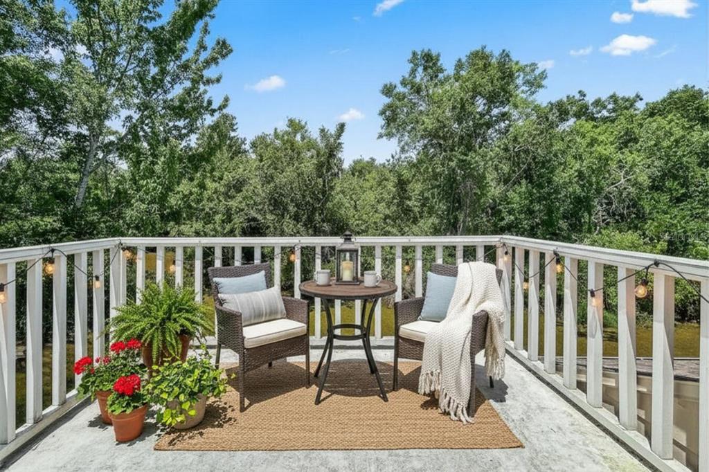 A balcony with a small table, two chairs, potted plants, and string lights overlooking trees — 6419 REMUS DRIVE, NEW PORT RICHEY, FL