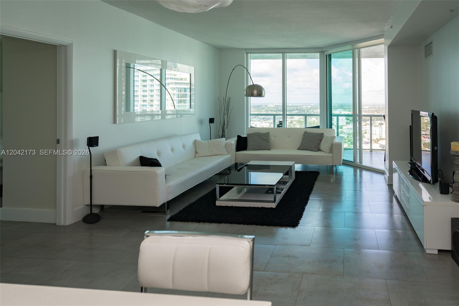 A modern living room features white leather sofas, a glass coffee table, and floor-to-ceiling windows — MIAMI, FL