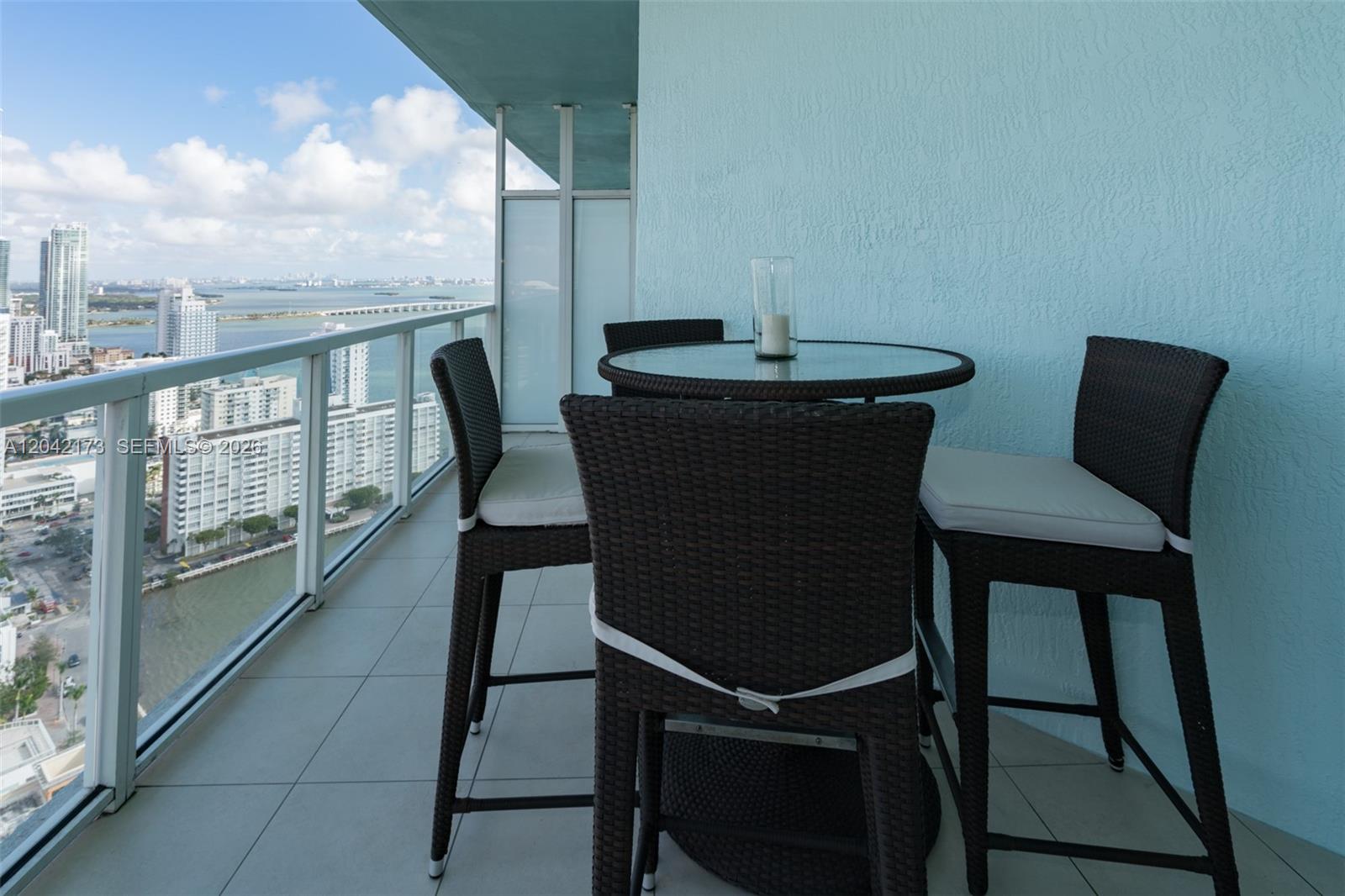 A balcony features a glass-topped table with four wicker chairs overlooking a city skyline — MIAMI, FL