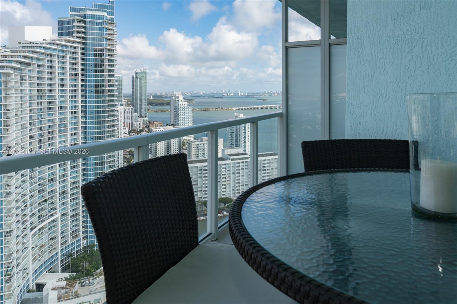 A high-rise balcony features a glass-topped table and chairs overlooking a city skyline and bay — MIAMI, FL
