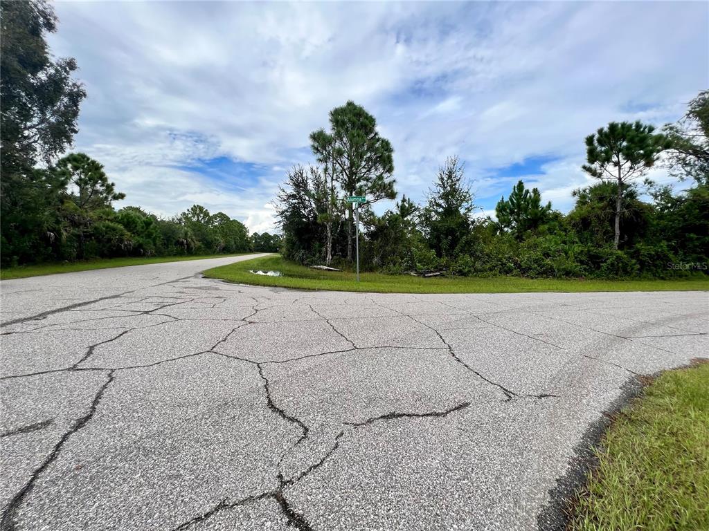 An asphalt road intersection is surrounded by dense green trees and vegetation under a cloudy sky — 12068 ALUMNA AVENUE, PORT CHARLOTTE, FL