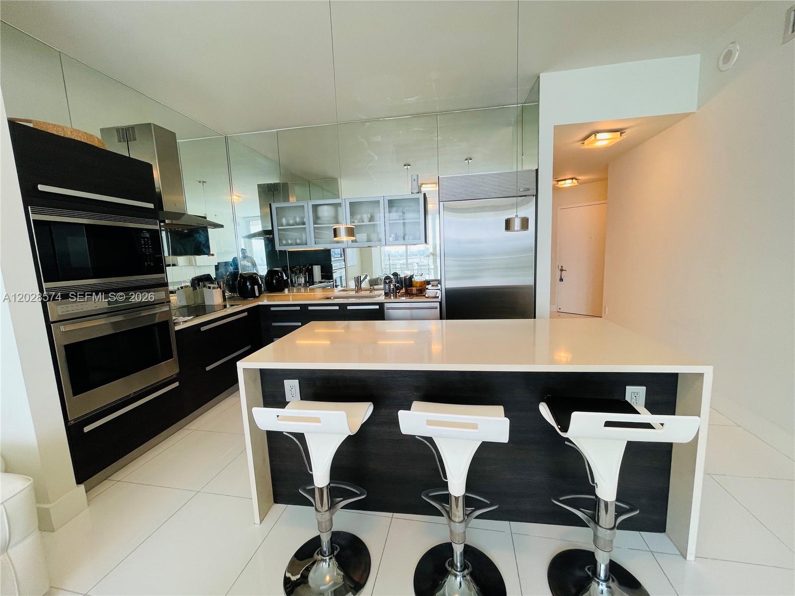 A modern kitchen features a white island with three stools, dark cabinetry, and mirrored walls — 2020 N Bayshore Dr # 2008, Miami FL 33137, MIAMI, FL