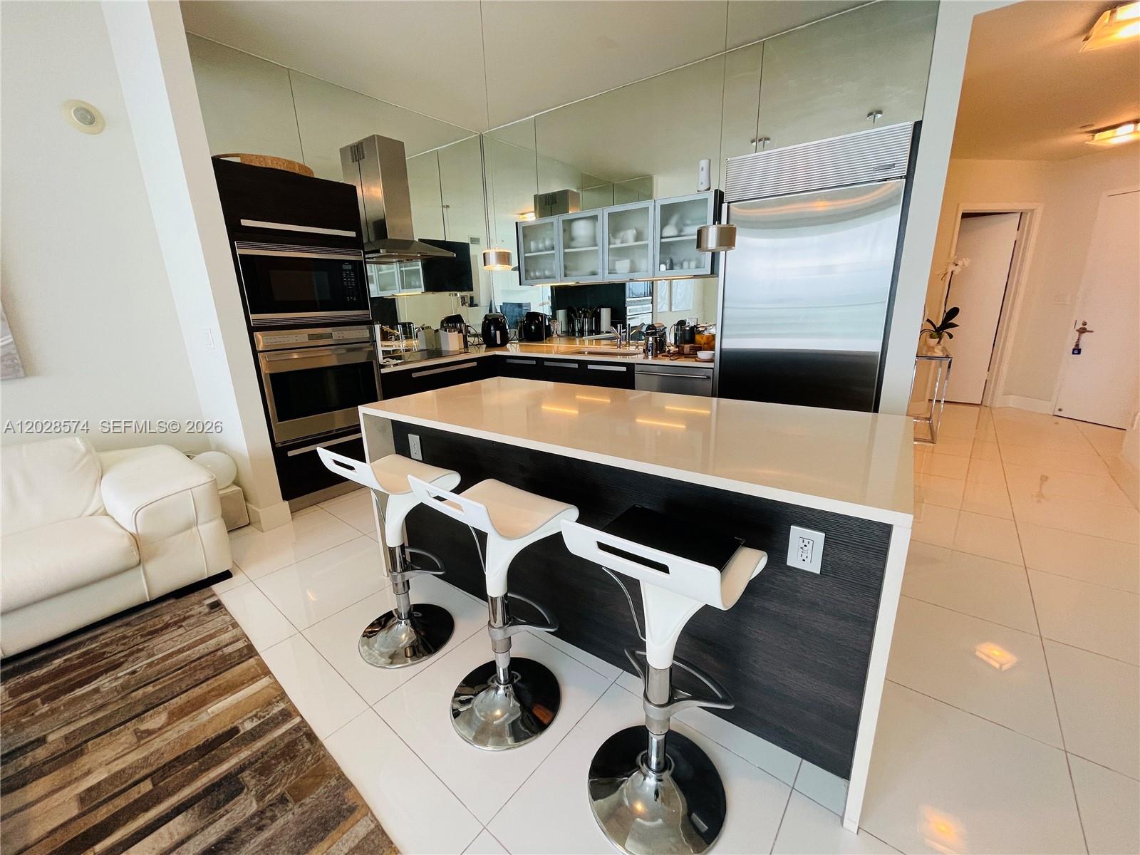 A modern kitchen features a white island with three stools, dark cabinetry, and mirrored walls — 2020 N Bayshore Dr # 2008, Miami FL 33137, MIAMI, FL