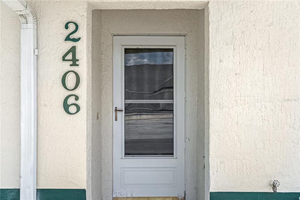 A white storm door is centered on a textured beige wall with vertical house numbers — 2406 KAM COURT, KISSIMMEE, FL