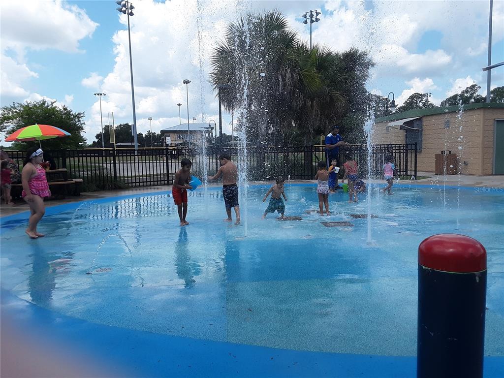 Children play in a public splash pad with water jets and a nearby building — KACHER ROAD, NORTH PORT, FL