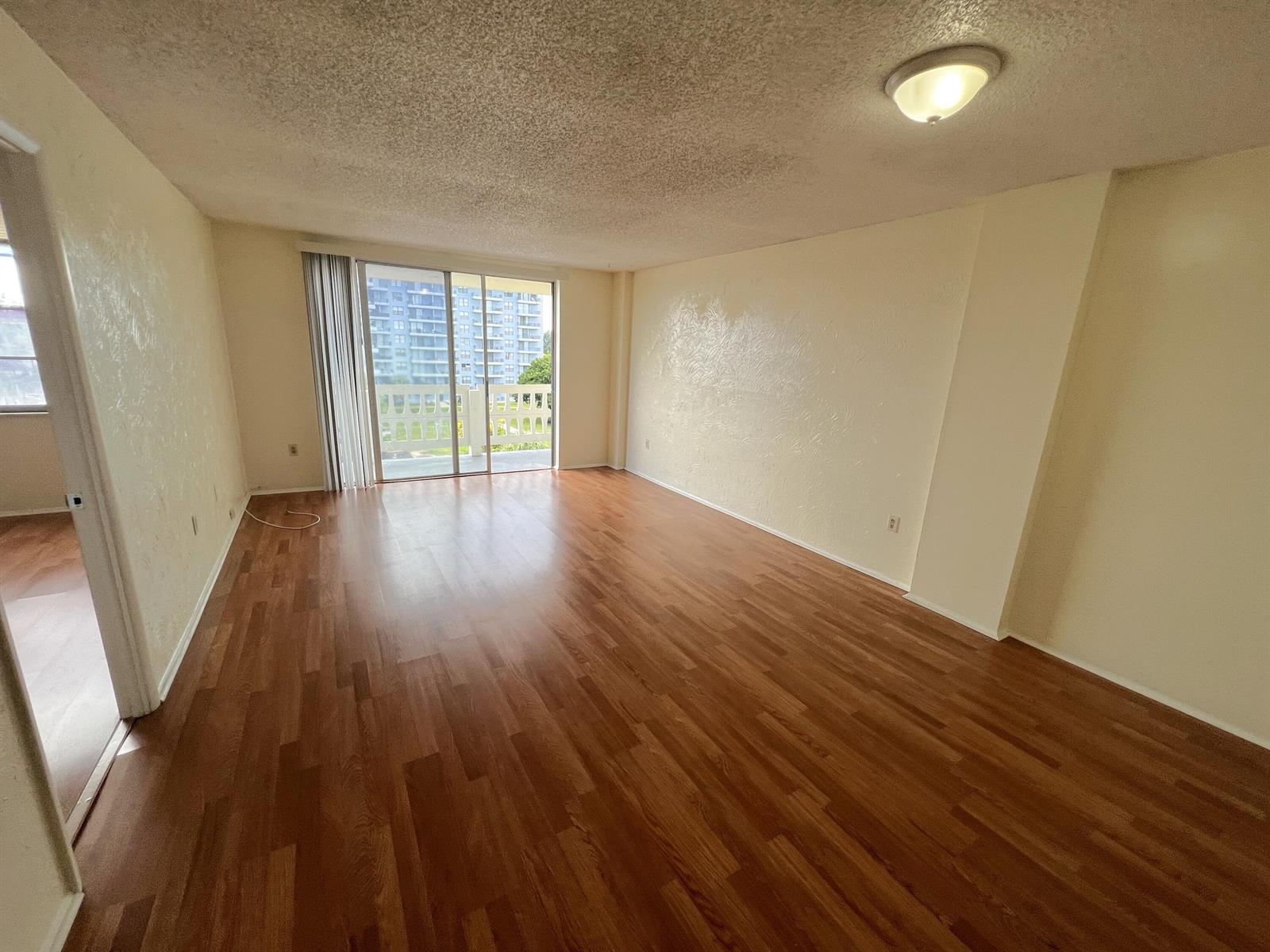An empty living room with wood-look flooring, textured walls, and a sliding glass balcony door — 15600 NW 7th Avenue # 516, Miami FL 33169, MIAMI, FL