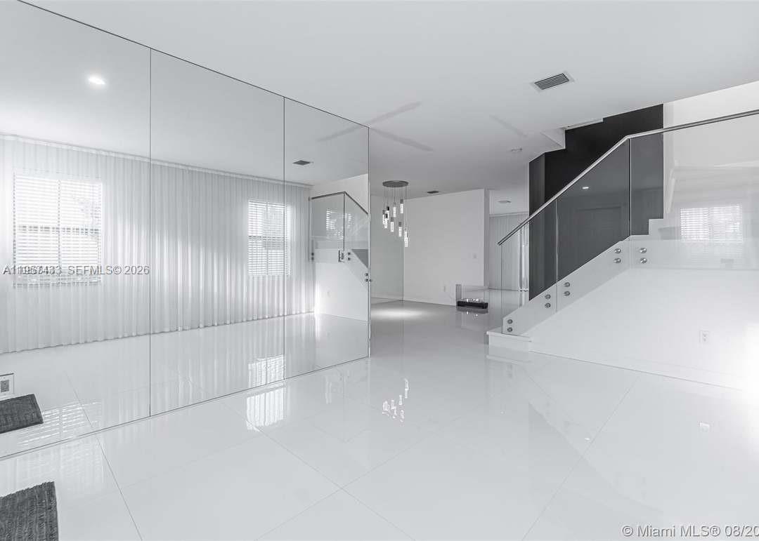 A modern, minimalist living area features white tile flooring, a mirrored wall, and a glass-railed staircase — 10301 W 32nd Ln, Hialeah FL 33018, HIALEAH, FL