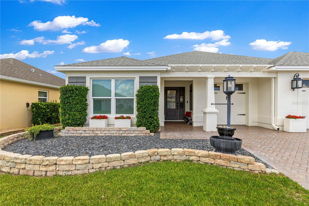 A single-story house exterior features a stone retaining wall, a paved driveway, and a fountain — 1132 MAYNARD PATH, THE VILLAGES, FL