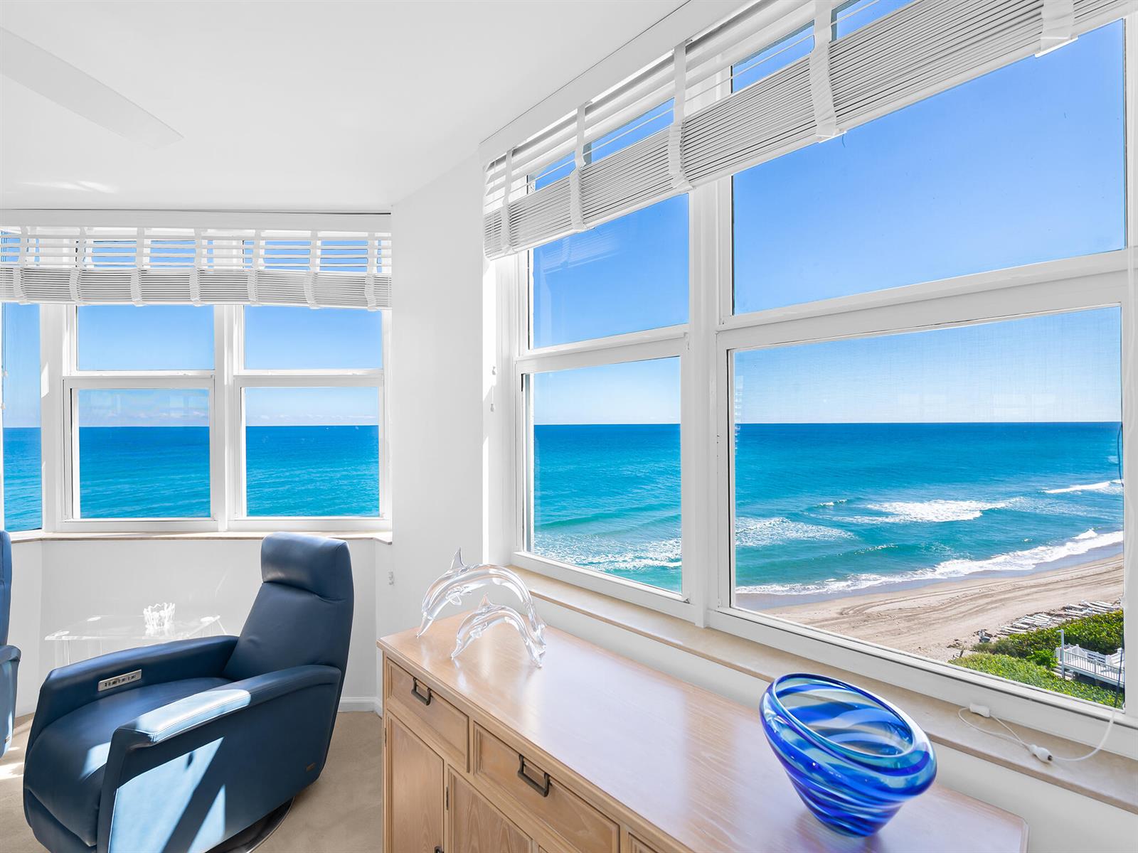 A room with a blue leather chair and wooden cabinet overlooks the ocean through windows — 3407 S Ocean Boulevard # 7a, Highland Beach FL 33487, HIGHLAND BEACH, FL