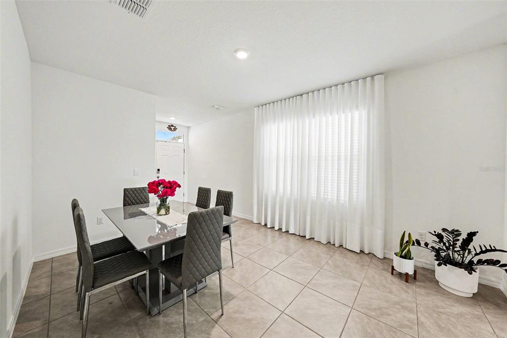 A dining room features a gray table, six chairs, tile flooring, and floor-to-ceiling white curtains — 6298 WANDERING WILLOW DRIVE, WESLEY CHAPEL, FL