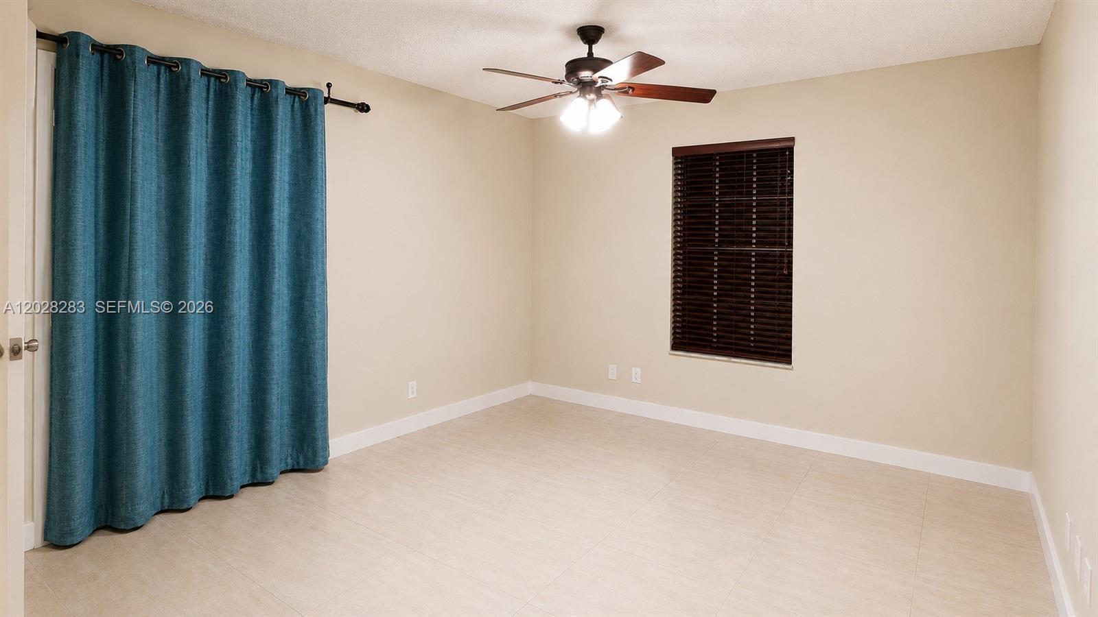 An empty room features beige walls, light tile flooring, a ceiling fan, and blue curtains — 6292 La Costa Dr # M, Boca Raton FL 33433, BOCA RATON, FL