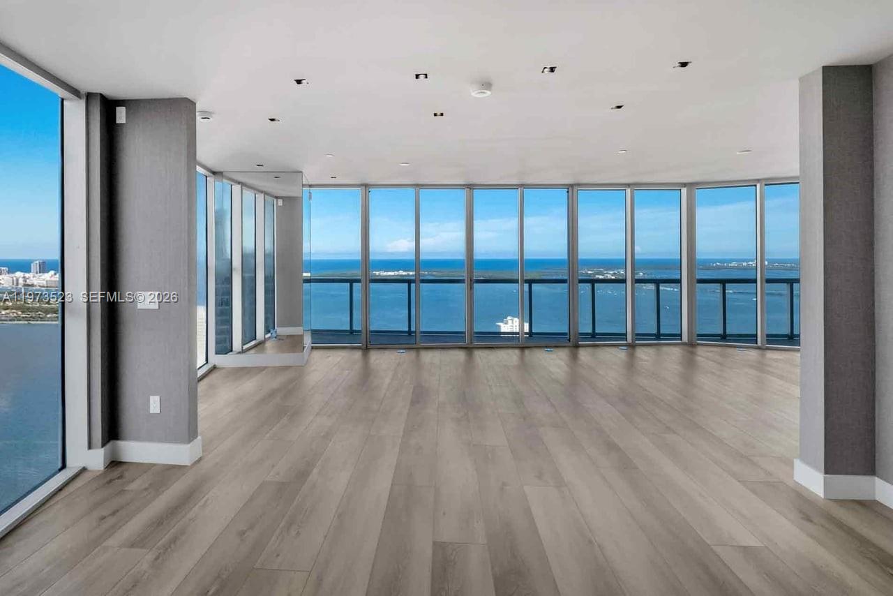 A spacious, empty room features light wood flooring, gray pillars, and floor-to-ceiling windows overlooking water — 465 Brickell Ave # 5701, Miami FL 33131, MIAMI, FL
