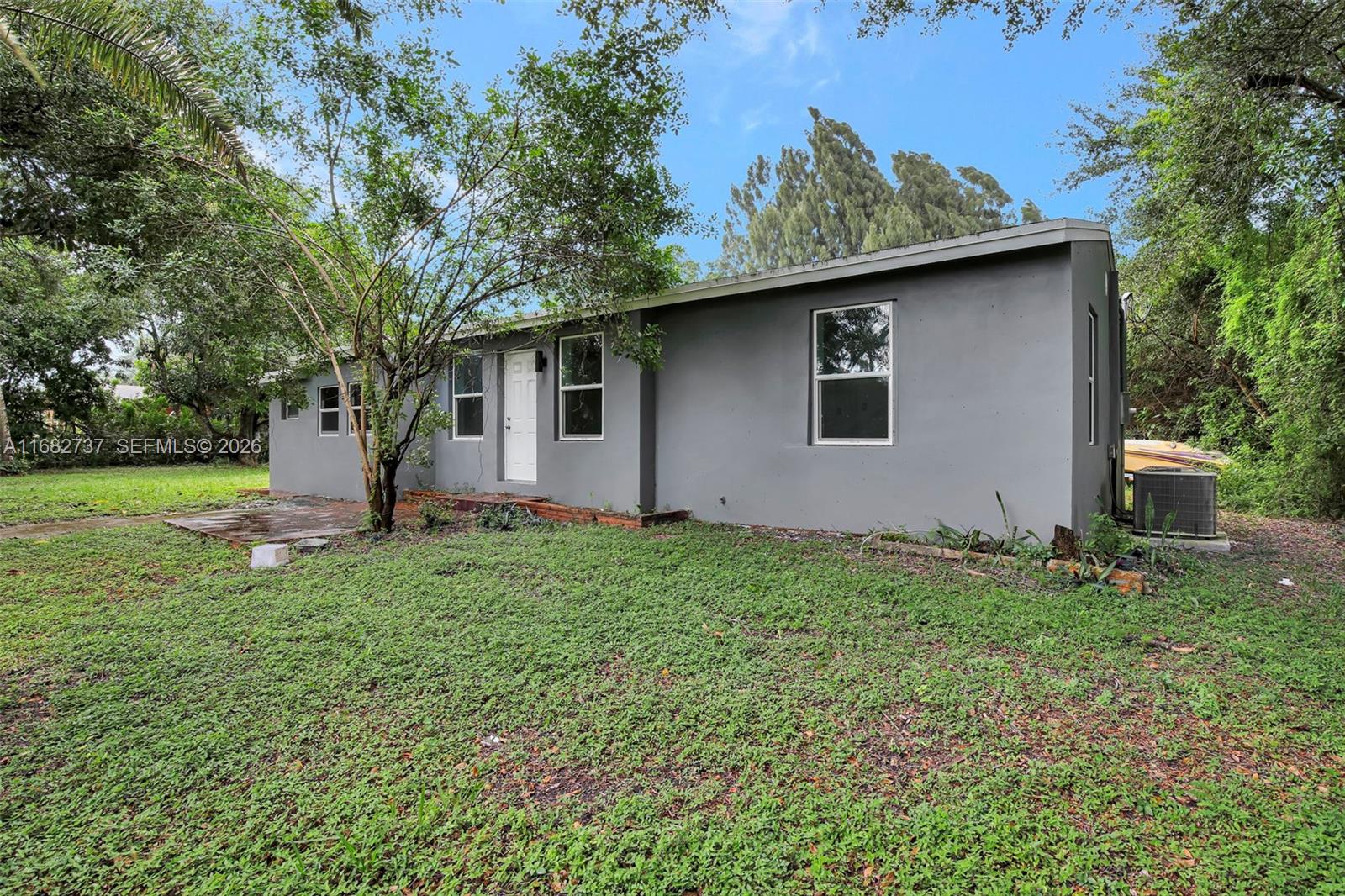 A single-story gray house with a white door stands on a grassy lot with trees — 4381 SW 51st St, Dania Beach FL 33314, DANIA BEACH, FL