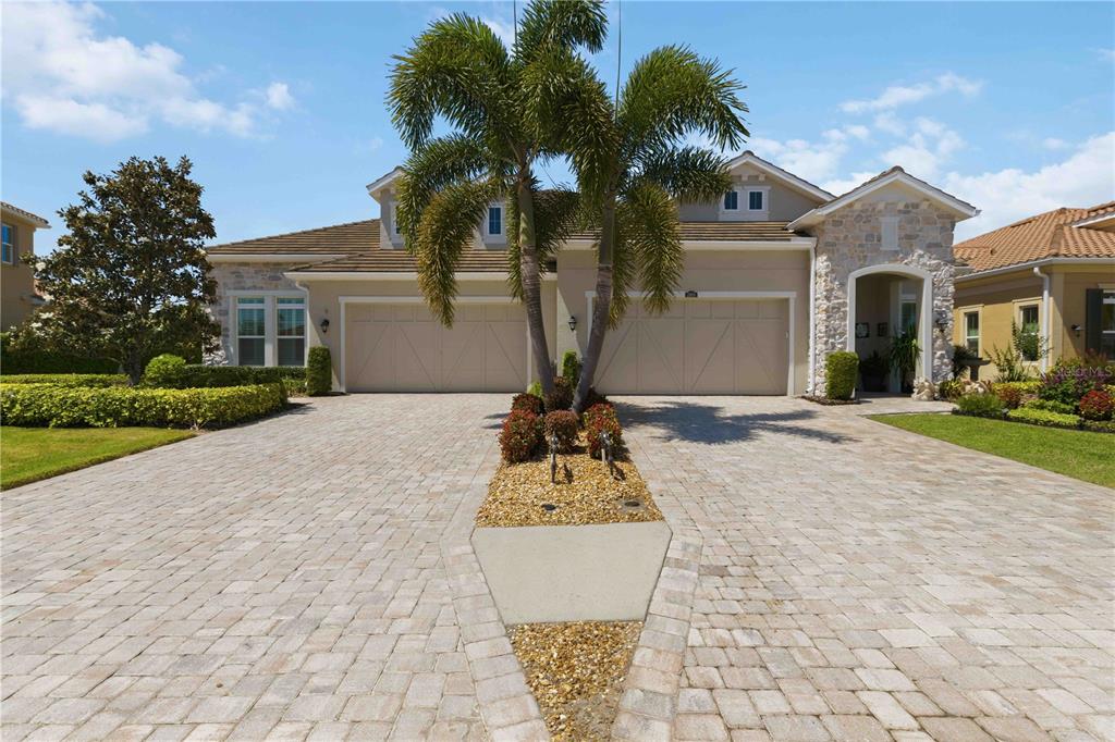 A beige house with a stone-accented entryway, three-car garage, and a palm tree-lined paver driveway — 2410 STARWOOD COURT, BRADENTON, FL