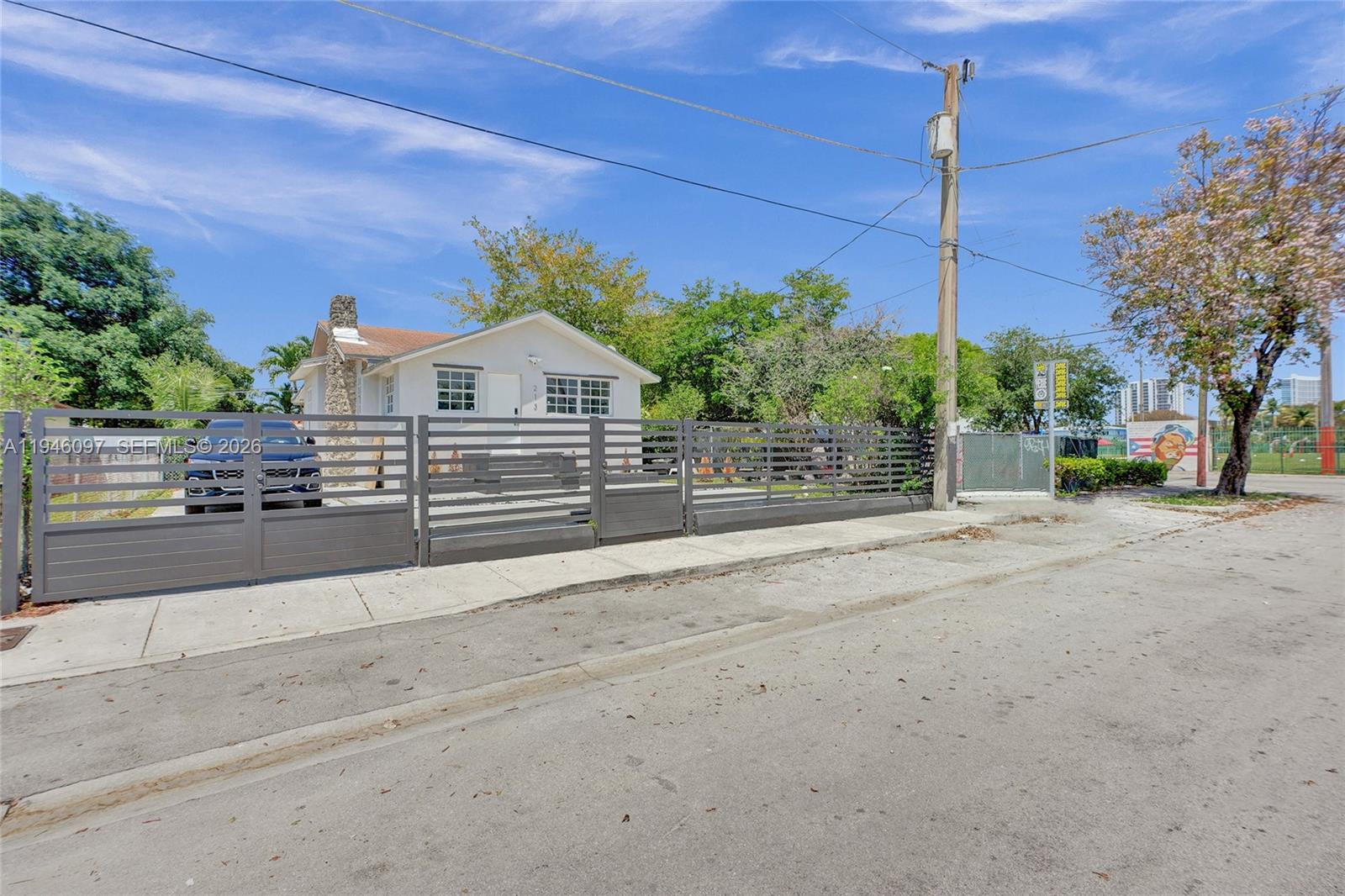 A single-story white house with a stone chimney sits behind a modern gray metal fence — 213 NW 34th Ter, Miami FL 33127, MIAMI, FL