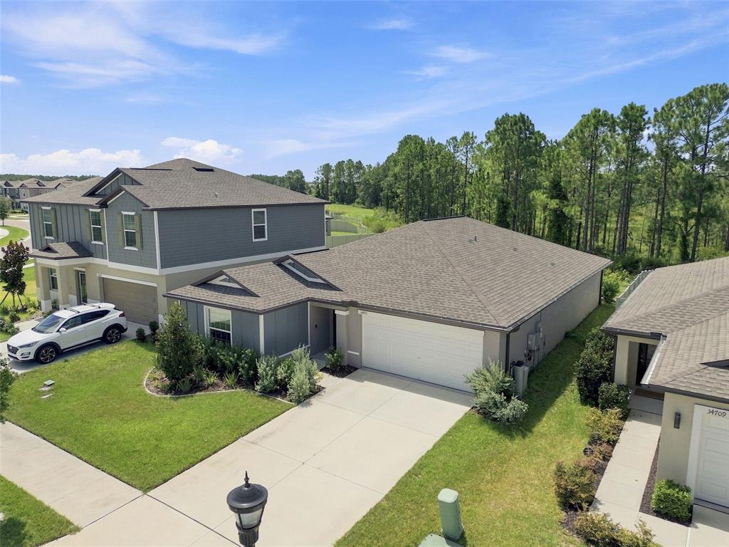 An aerial view shows a gray suburban house with a two-car garage and green lawn — 34701 RAINVILLE LOOP, ZEPHYRHILLS, FL