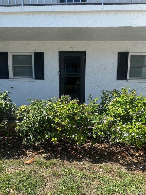 A white exterior wall features a dark front door numbered 207 between two shuttered windows — 202 46TH TERRACE W UNIT #207, BRADENTON, FL