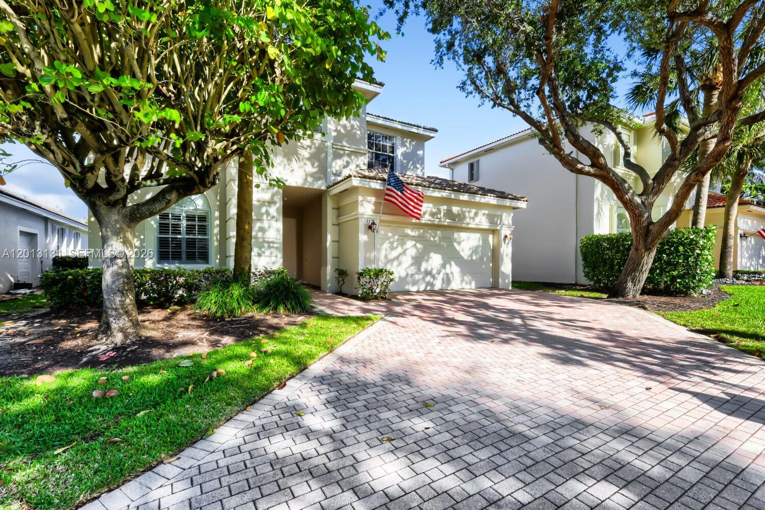 A two-story house features a brick driveway, a garage, and large trees in the front — 117 Palm Cir, Atlantis FL 33462, ATLANTIS, FL