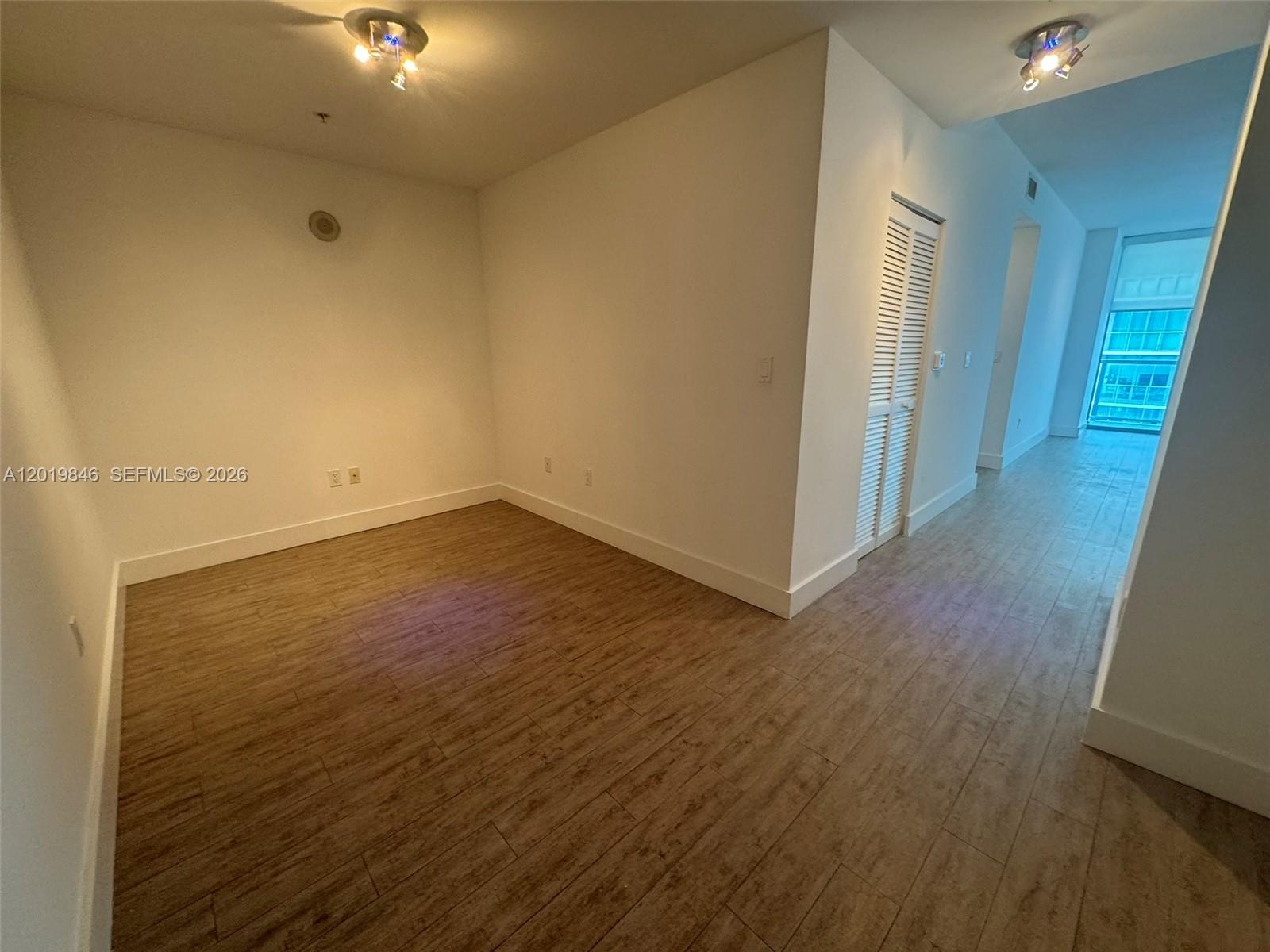 An empty room with light wood-look tile flooring and white walls leads to a hallway — 79 SW 12th St, Miami FL 33130, MIAMI, FL
