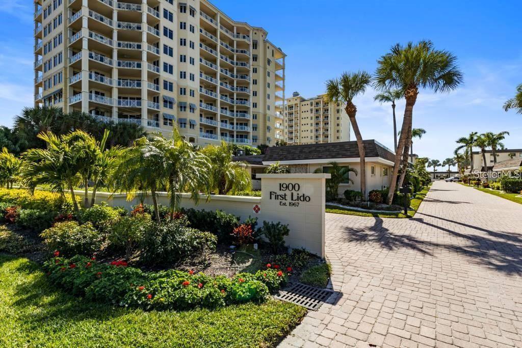 A paved driveway leads to a multi-story apartment building with a sign reading First Lido — 1900 BENJAMIN FRANKLIN DRIVE UNIT V-15, SARASOTA, FL