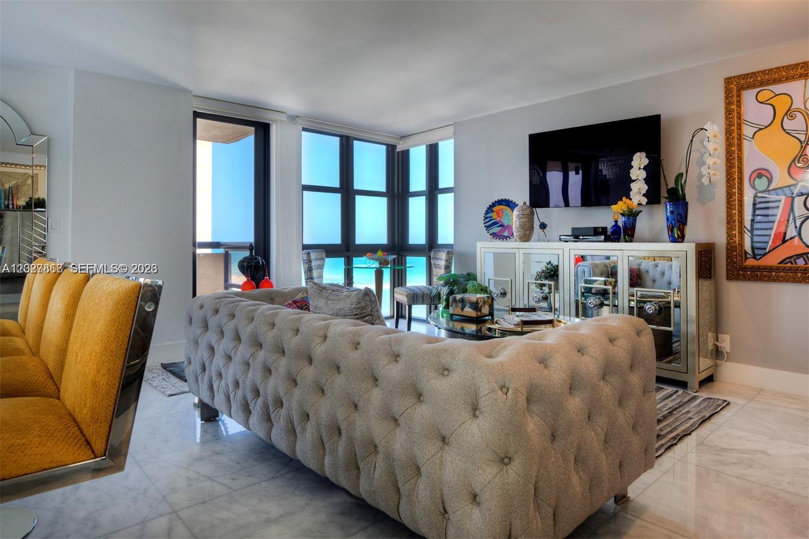 A living room features a tufted sofa, mirrored console, and floor-to-ceiling windows overlooking the ocean — 2555 Collins Ave # 1400, Miami Beach FL 33140, MIAMI BEACH, FL