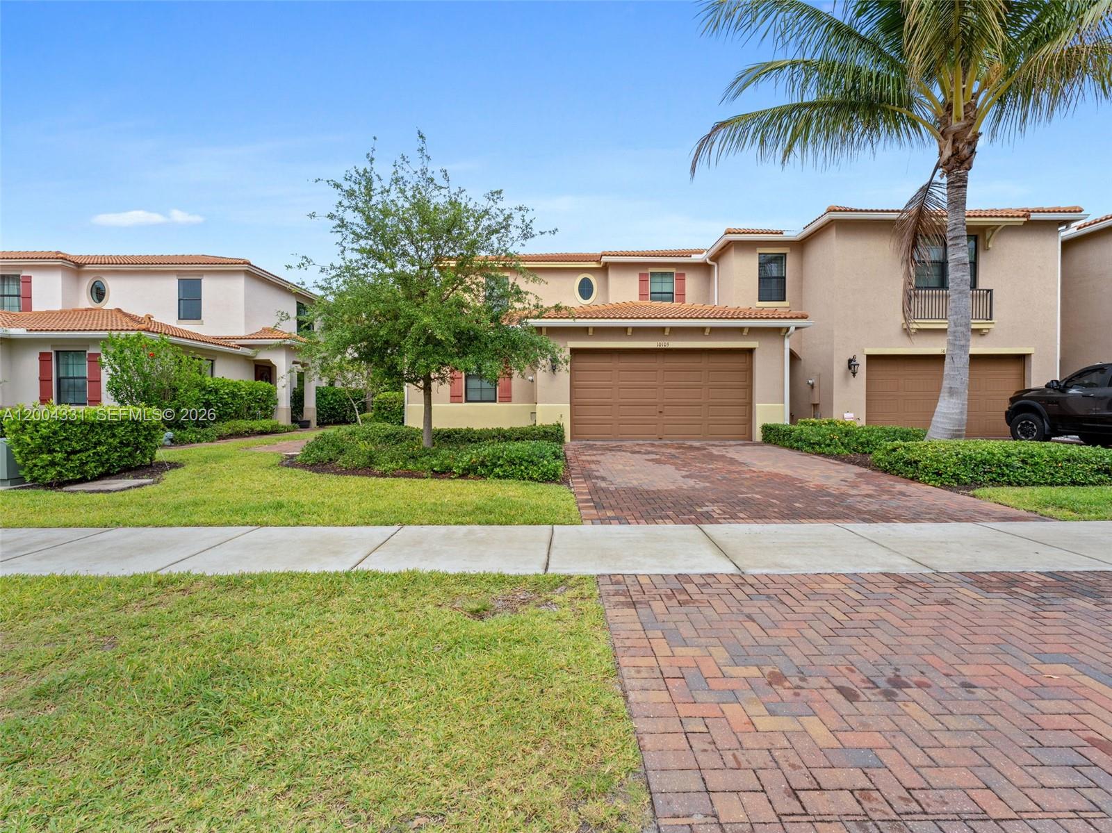 A two-story stucco townhouse features a brown garage door, brick driveway, and a palm tree — 10105 Brickhill Dr, Boca Raton FL 33428, BOCA RATON, FL