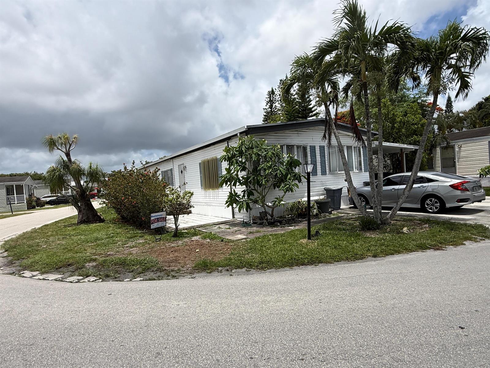 A white mobile home with a for-sale sign sits on a corner lot with palm trees — 4707 Nancy Drive # 757, FL 33463, FL