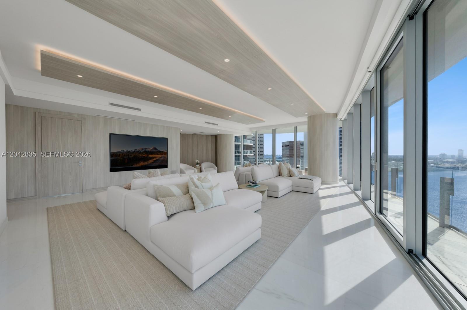 A modern living room features white sectionals, light wood paneling, and floor-to-ceiling windows overlooking water — 300 Biscayne Boulevard Way # 2701, Miami FL 33131, MIAMI, FL