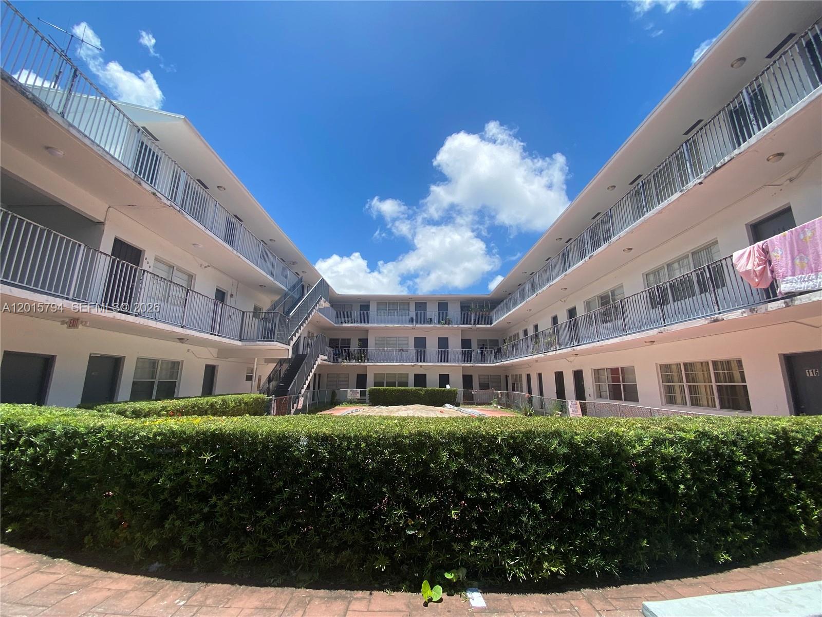A courtyard view of a multi-story apartment complex with exterior walkways and a central hedge — 13700 NE 6th Ave # 115, North Miami FL 33161, NORTH MIAMI, FL
