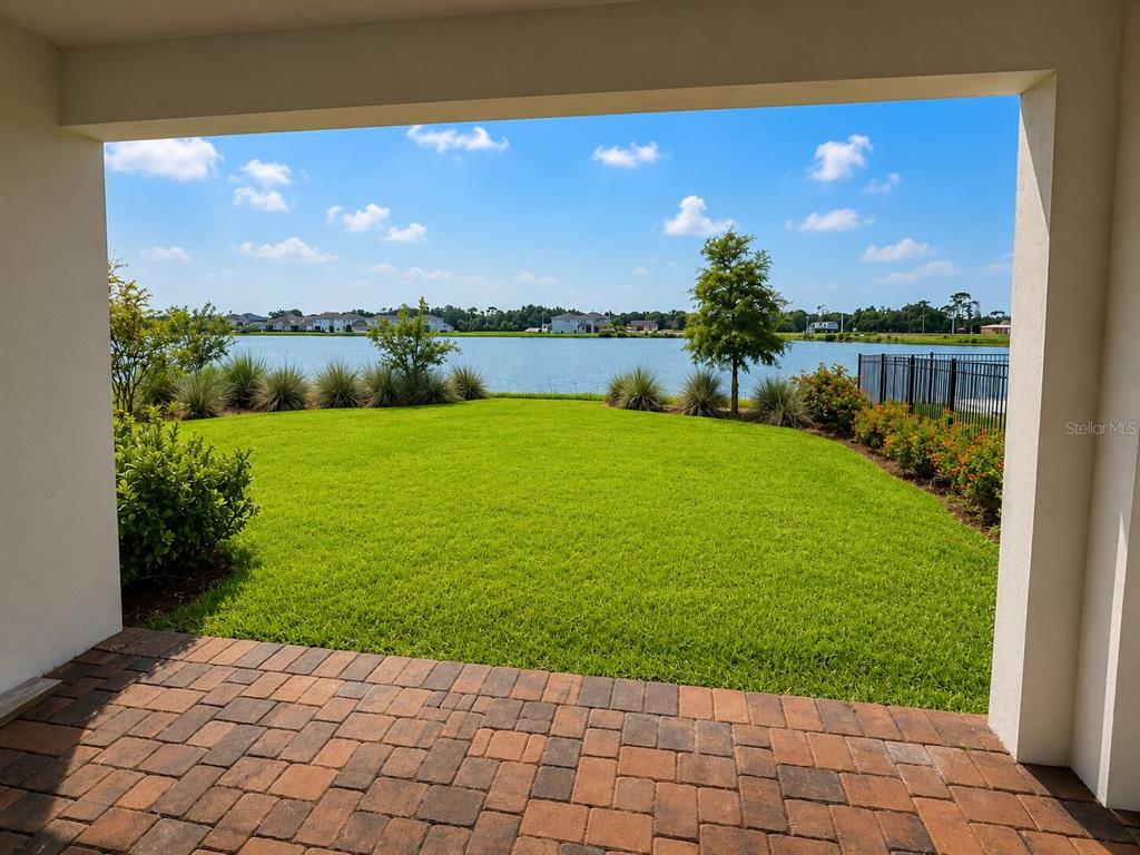 A covered brick patio overlooks a grassy backyard and a lake under a blue sky — 13629 MCMILLAN DRIVE, ORLANDO, FL