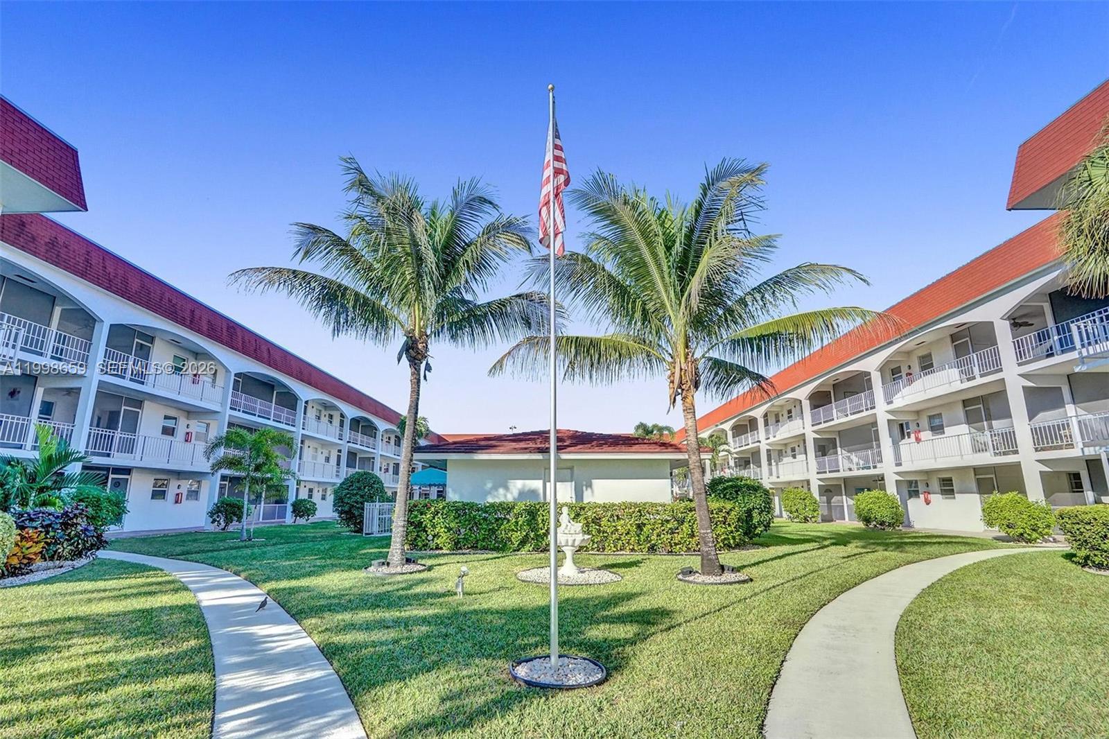 A courtyard with a flagpole, palm trees, and walkways between two multi-story apartment buildings — 580 Egret Dr # 212, Hallandale Beach FL 33009, HALLANDALE BEACH, FL