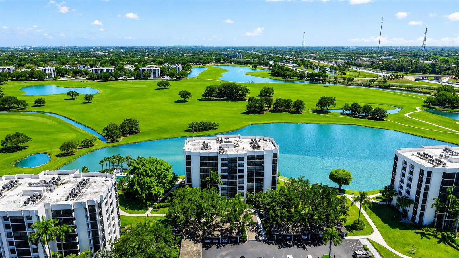 An aerial view shows three white apartment buildings situated beside a large golf course lake — 7768 Lakeside Boulevard # 551, Boca Raton FL 33434, BOCA RATON, FL