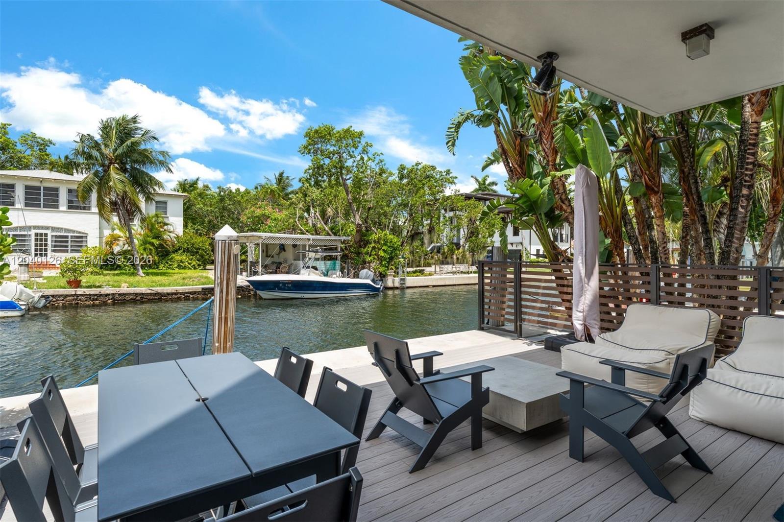 A wooden deck features outdoor dining and lounge furniture overlooking a canal with a boat — 3560 W Fairview St, Miami FL 33133, MIAMI, FL