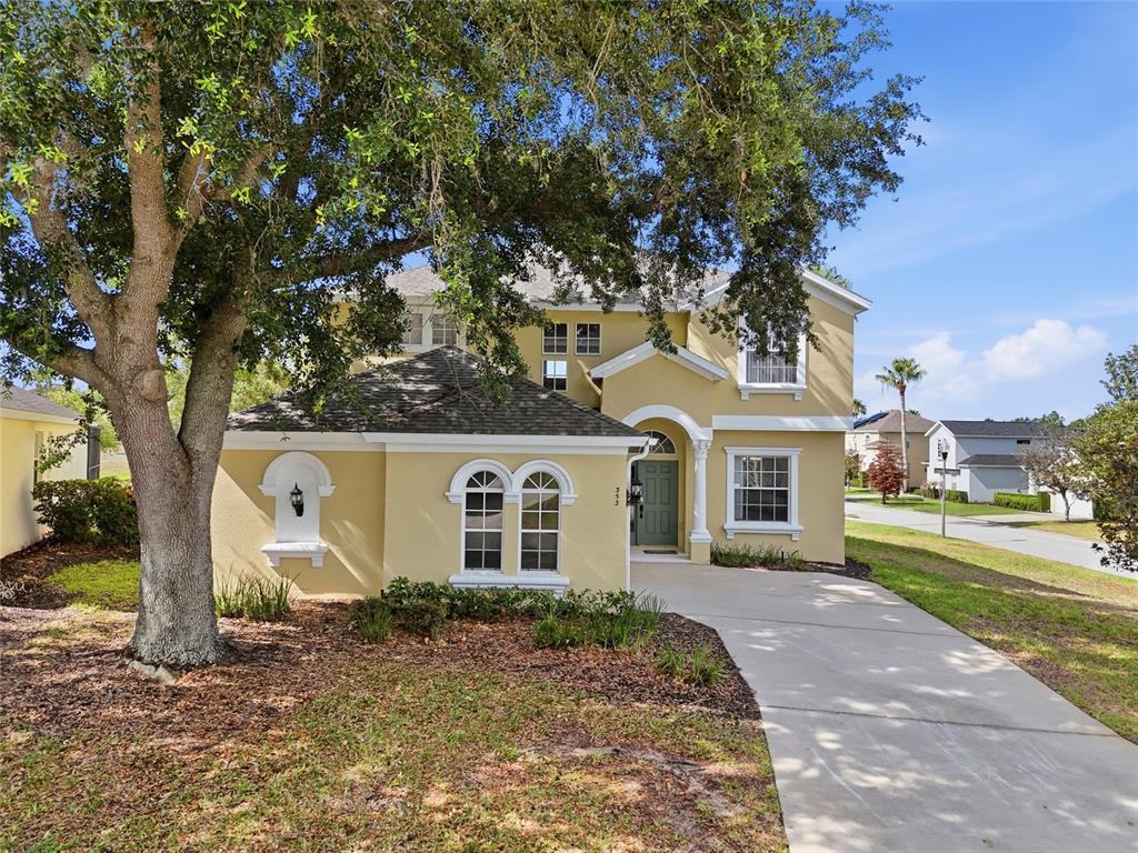 A two-story yellow house features a concrete driveway, a large tree, and arched windows — 353 PLUMOSO LOOP, DAVENPORT, FL