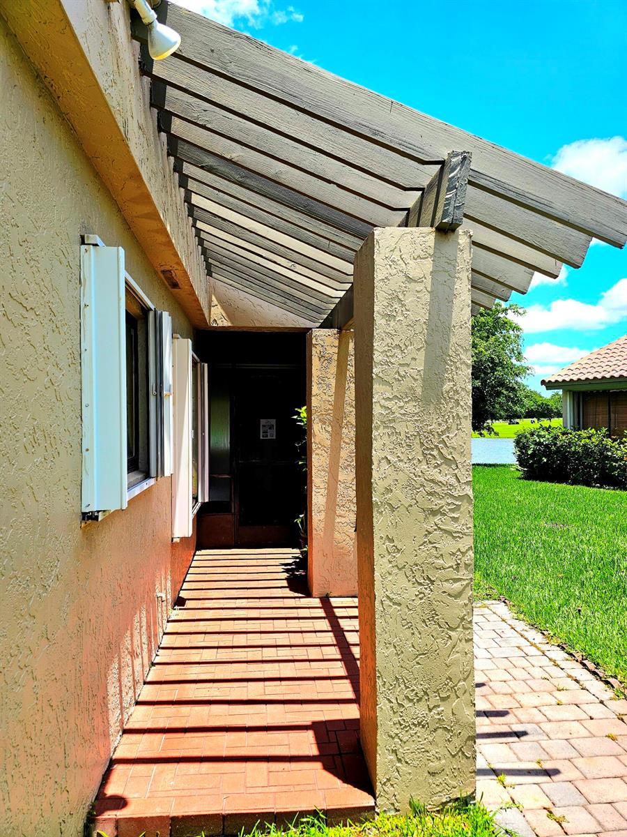 A covered walkway with a wooden pergola leads to a front entrance beside a stucco wall — 566 Village Lake Drive, Weston FL 33326, WESTON, FL