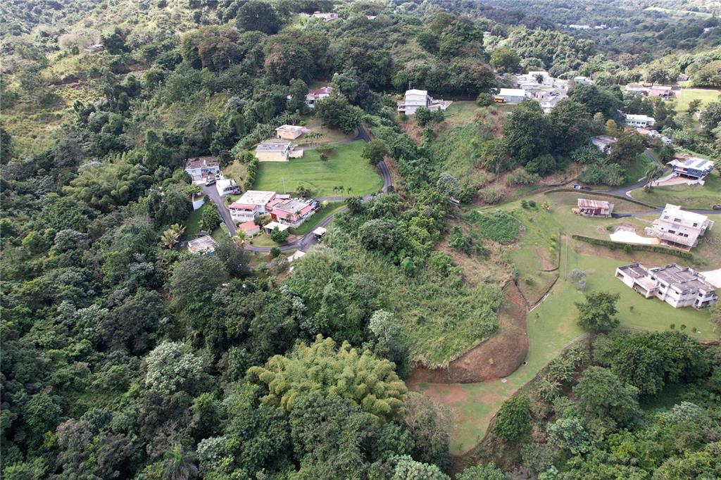 An aerial view shows several houses scattered across a hilly, forested landscape with grassy lawns — PR 568 KM 28.5 SECTOR EL LIMON PADILLA, COROZAL, PR