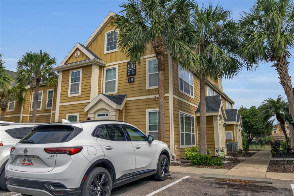 A white SUV is parked in front of a three-story yellow apartment building with palm trees — 5511 ROSEHILL ROAD UNIT 203, SARASOTA, FL