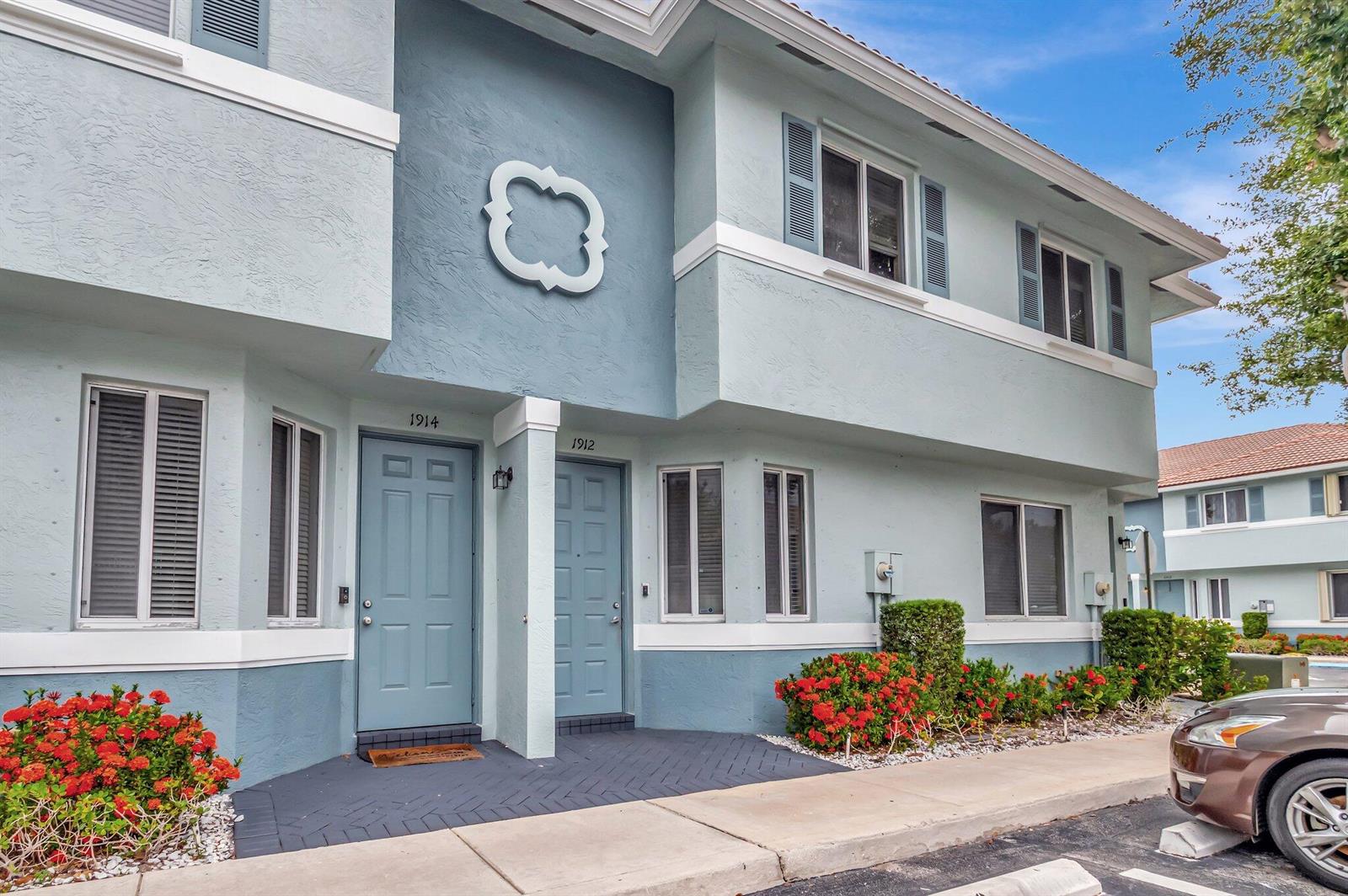 A two-story building exterior features light blue stucco walls, two front doors, and red flowers — 1912 Hibiscus Lane, Riviera Beach FL 33404, RIVIERA BEACH, FL