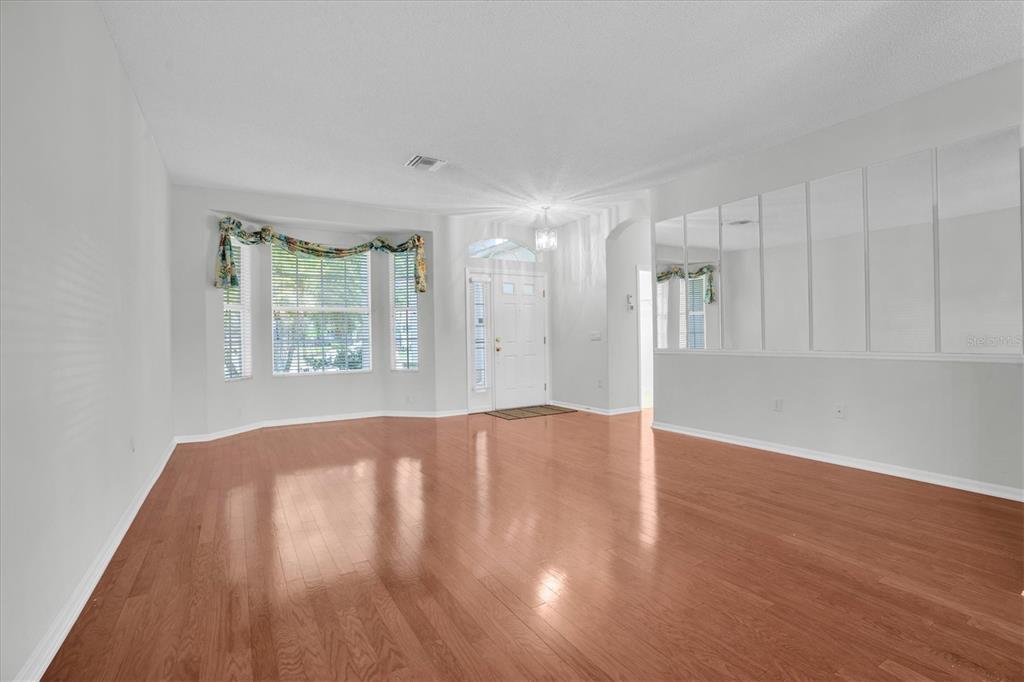 An empty living room features light-colored walls, hardwood flooring, a bay window, and mirrored wall panels — 1510 MORNING ROSE PLACE, TRINITY, FL