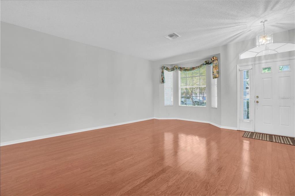 An empty living room features light wood flooring, a bay window, and a white door — 1510 MORNING ROSE PLACE, TRINITY, FL
