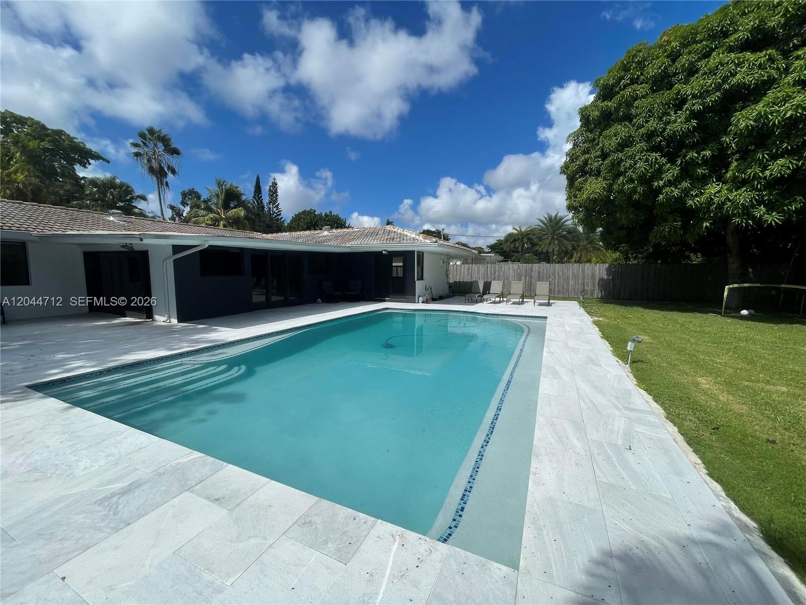 A rectangular swimming pool sits on a stone patio next to a house and lawn — 2115 Alamanda Dr, North Miami FL 33181, NORTH MIAMI, FL