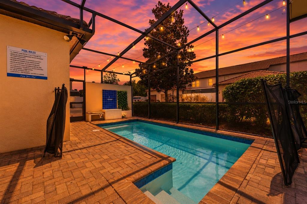 A screened-in patio features a rectangular swimming pool, brick pavers, and string lights at sunset — 1974 MAJORCA DRIVE, KISSIMMEE, FL