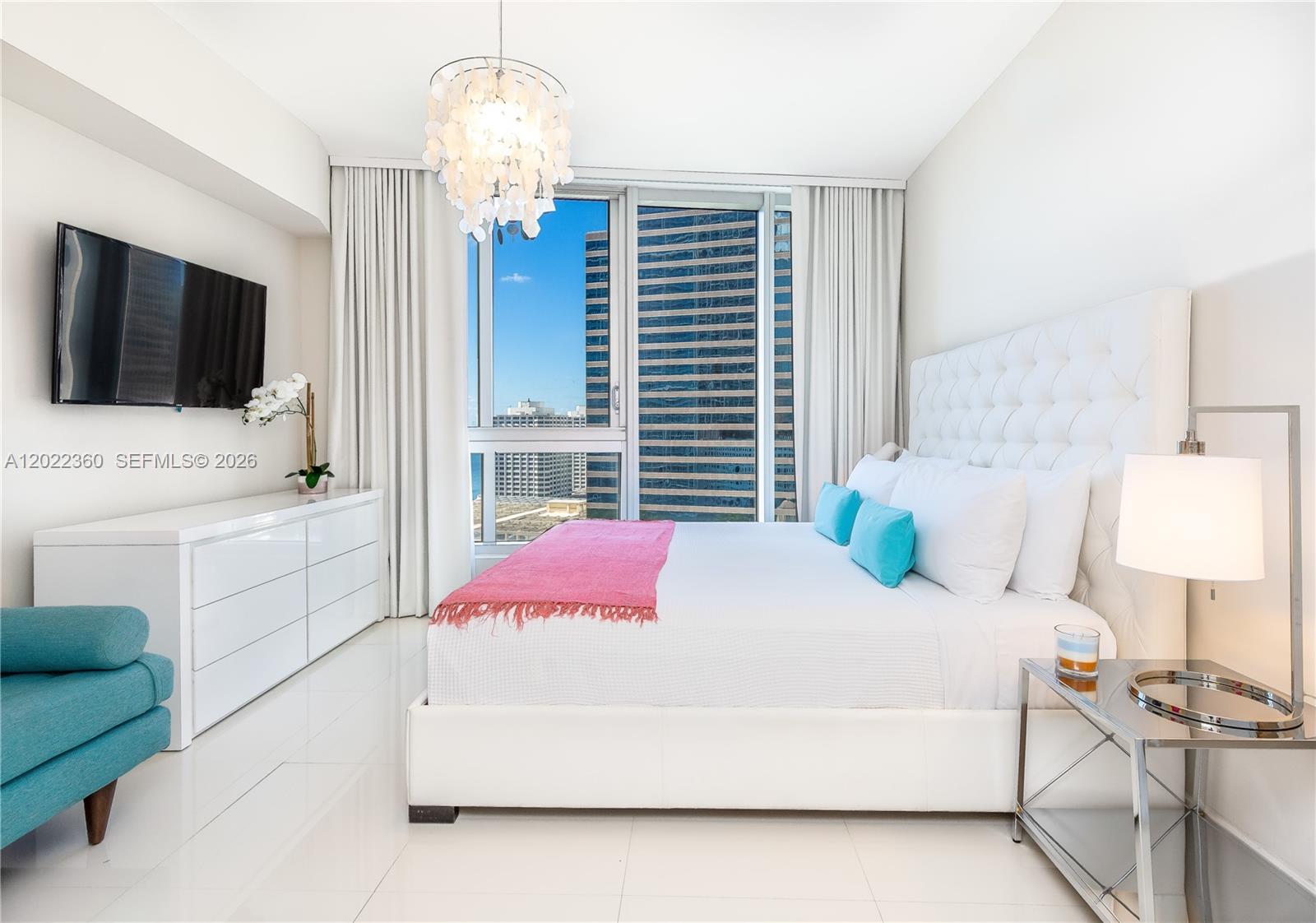 A modern bedroom features a white tufted bed, a dresser, and floor-to-ceiling city views — MIAMI, FL