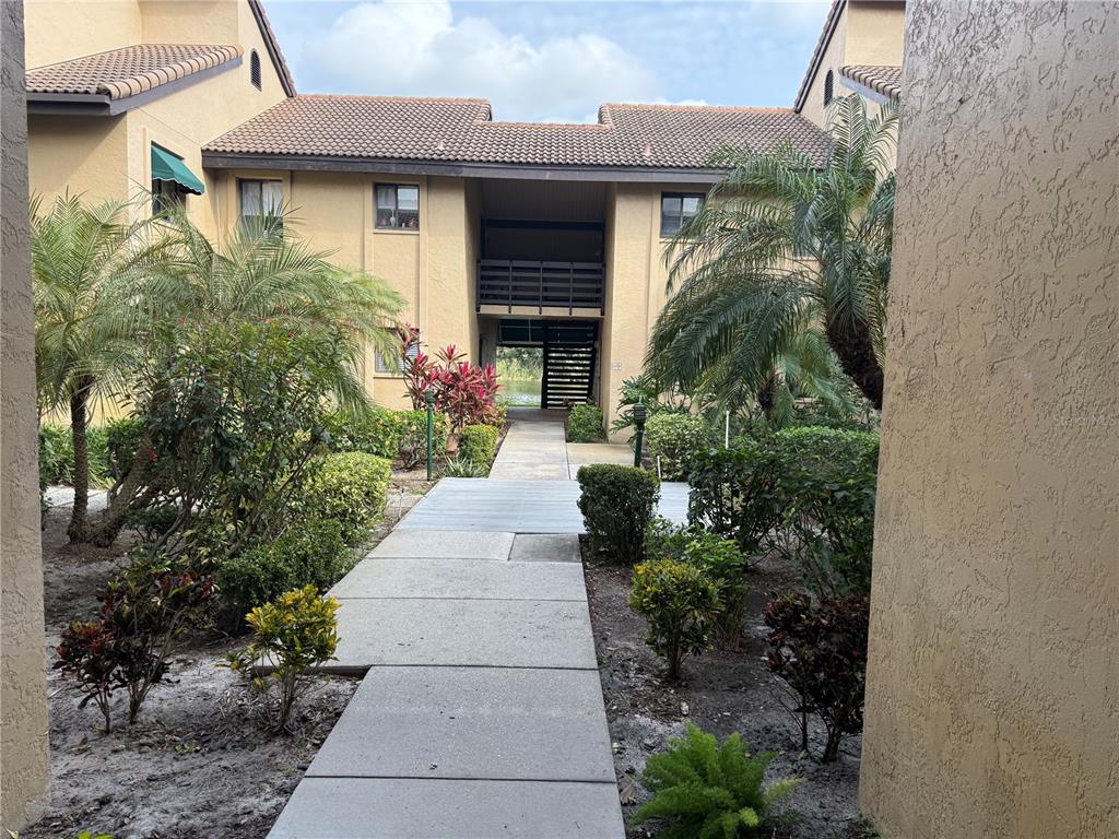 A concrete walkway leads through a landscaped courtyard toward a two-story tan apartment building — 5631 ASHTON LAKE DRIVE UNIT 7, SARASOTA, FL