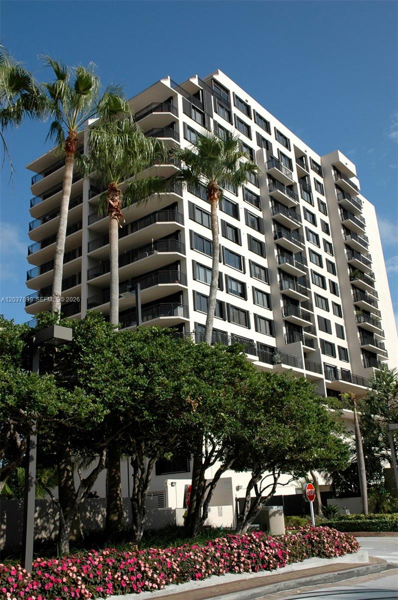 A tall, multi-story residential building with balconies stands behind palm trees and flowering bushes — 540 Brickell Key Dr # 1513, Miami FL 33131, MIAMI, FL