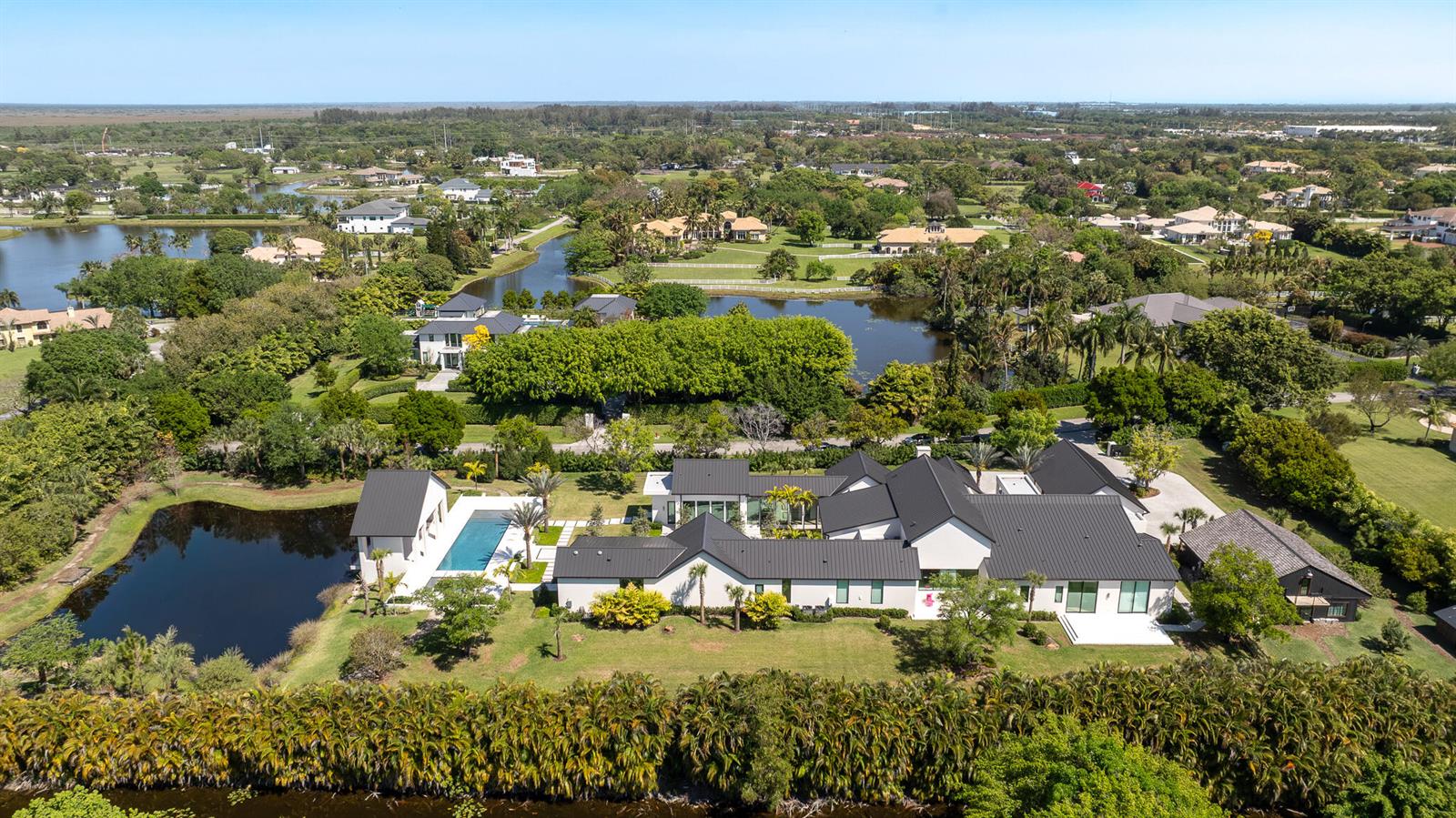 An aerial view shows a large modern house with a black roof, pool, and pond — 10468 El Caballo Court, Delray Beach FL 33446, DELRAY BEACH, FL