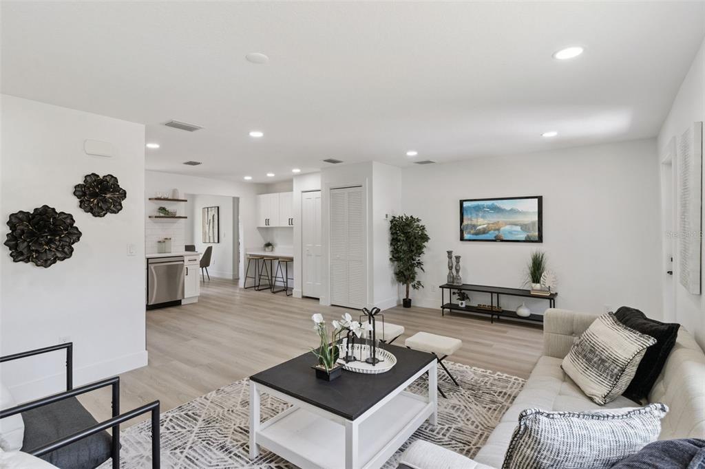 A bright, open-concept living room features light wood flooring, a modern kitchen, and neutral furniture — 7211 ORPINE DRIVE N, ST PETERSBURG, FL